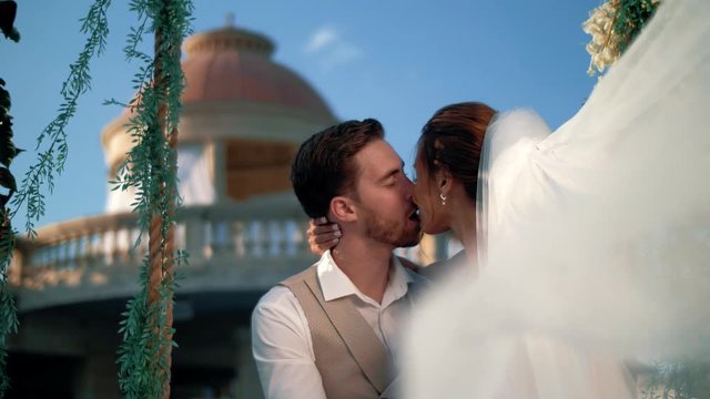 Beautiful newlyweds kiss while sitting on a swing in a Greek antique location