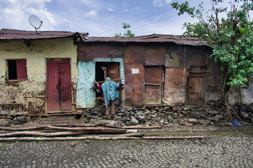 A traveling chicken seller with a pair of chickens hanging from a wooden stick around his neck next to a humble, rusty, painted metal house. Africa.