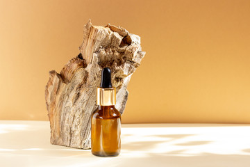 A glass cosmetic bottle with a dropper stands next to a bar of wood on a beige background with...