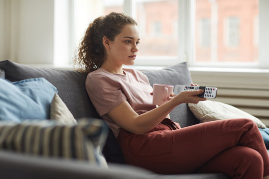 Side view portrait of young woman watching TV and holding remote while sitting on sofa at home in cozy apartment, copy space
