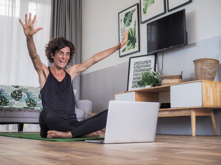 happy excited and smiling women sitting crossed legged on the floor on a yoga mat in her living room at home looking at laptop having her hands up celebrating