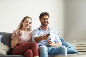 Obraz premium Portrait of modern couple watching TV and holding mugs while sitting on sofa at home in cozy apartment enjoying lazy time, copy space