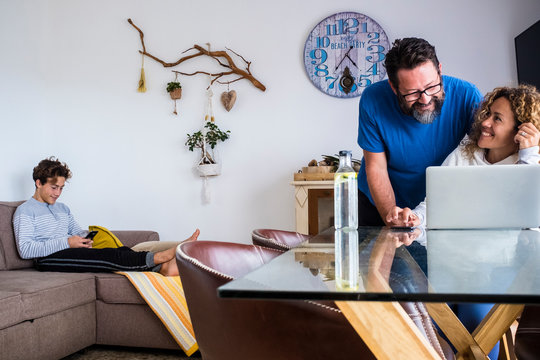 Caucasian Family At Home Lifestyle With Technology - Couple Husband And Wife Use Laptop Computer On The Table And Teenager Son With Phone On The Couch - Quarantine Lockdown Concept