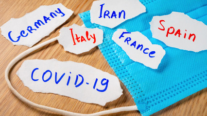 Coronavirus COVID-19 inscription on paper near medical face mask on yellow table. Italy, France, Spain, Germany and Iran. Novel pneumonia 2019-nCoV. Wuhan's seafood market virus