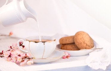 White porcelain cup of tea, milk jug, and fresh-baked oat cookies. English breakfast still-life with drink and treats and tablecloth.