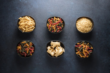 6 bowls of raw pasta, noodles, macaroni, noodles and colored curls on a rustic table