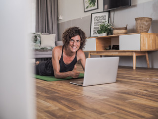 women lying on the floor on her stomach on a green mat in her living room at home looking at laptop screen thumb up getting ready to do an online sport / yoga session