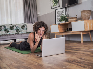 women lying on the floor on her stomach on a green mat in her living room at home looking at laptop and waving into the screen getting ready to do an online sport / yoga session - Powered by Adobe