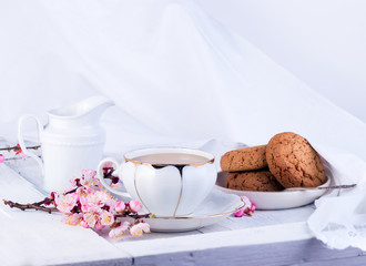 White porcelain cup of tea, milk jug, and fresh-baked oat cookies. English breakfast still-life with drink and treats and tablecloth.