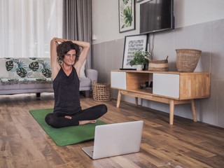 happy and smiling woman stretching her arm and looking into the laptop in doing a yoga pilates workout in her living room at home