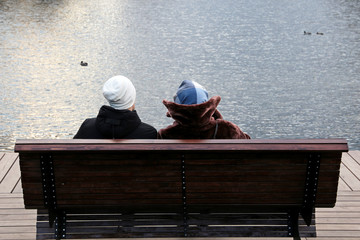 Couple in warm clothes sitting on a wooden bench on the shore of a lake with mallard ducks. Spring leisure in a park