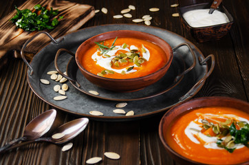 Clay dish with homemade rustic pumpkin soup with seeds on metal tray with seeds and greens aside