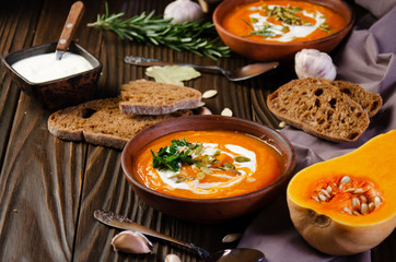Homemade rustic pumpkin soup with seeds in clay dish on wooden table with bread spices and greens aside