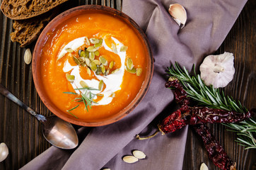Flat lay view at homemade rustic pumpkin soup with seeds in clay dish on wooden table with bread spices and greens aside