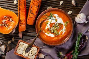 Flat lay view at homemade rustic pumpkin soup with seeds in clay dish on wooden table with bread spices and greens aside