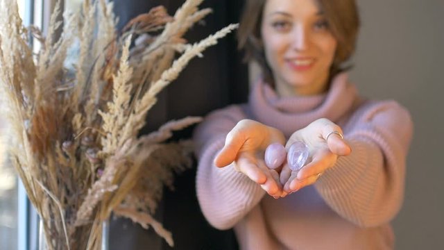 Female hands holding two yoni eggs made from pink quartz and transparent violet amethyst for vumfit, imbuilding or meditation near the window with vase of spikelets.
