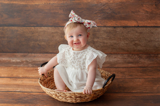 Portrait Of The Cute One Year Old Girl. With Headband And In White Dress. On Wooden Background. Caucasian Toddler Little Baby. With First Teeth. 