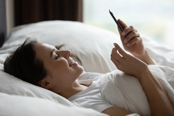 Smiling young woman lying in white bed wake up texting messaging on cellphone device, happy...