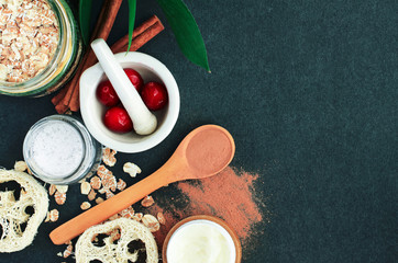 Homemade skin care treatment, beauty spa vegan ingredients top view dark grey background. 
Red cranberry in bowl, cinnamon powder, oatmeal flakes for face and body exfoliation cleansing scrub.
