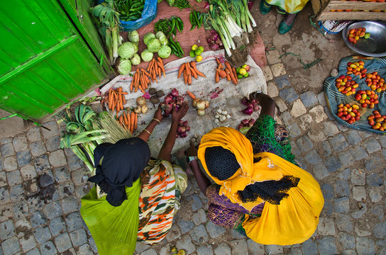Black women in colorful traditional African costumes crouching at a vegetable stand on the ground and buying vegetables. Take a zenith. Carrots, tomatoes, onions, garlic, leeks, peppers.