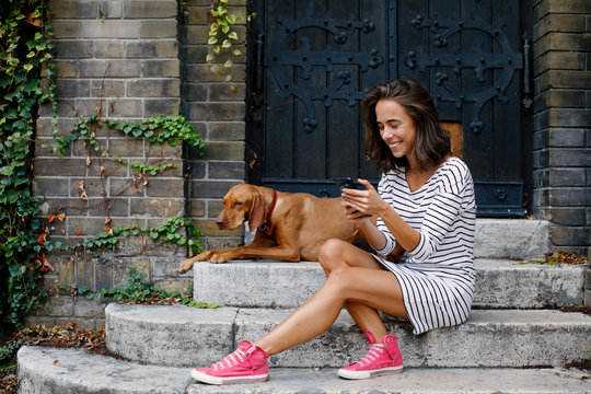 Young Woman Using Smartphone With Her Dog In The Backyard