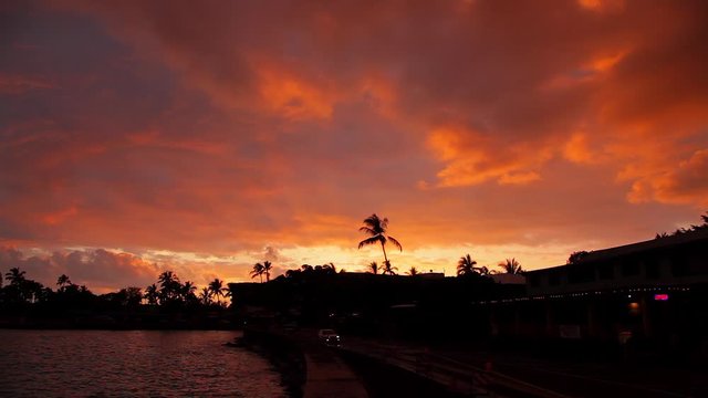 Brilliant red sunset in Kona on the island of Hawaii with a couple cars with only their white headlights seen slowly driving by a row of businesses.