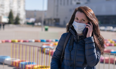 Fototapeta premium a beautiful girl in a jacket and a medical anti virus mask is talking on the phone in the square against the background of shopping centers and a Playground