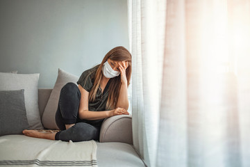 Sad lonely girl isolated stay at home in protective sterile medical mask on face looking at window,...