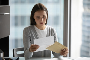 Pensive young woman sit at desk in office open envelope read post correspondence, focused...