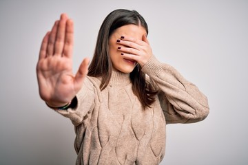 Young beautiful brunette woman wearing casual sweater standing over white background covering eyes with hands and doing stop gesture with sad and fear expression. Embarrassed and negative concept.