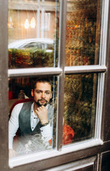 Portrait of bearded man in gray waistcoat sitting outside window in restaurant. View from the yard