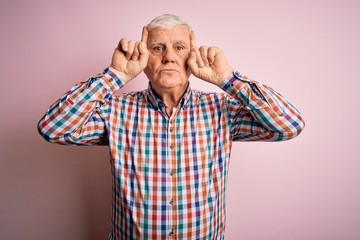 Senior handsome hoary man wearing casual colorful shirt over isolated pink background doing funny gesture with finger over head as bull horns