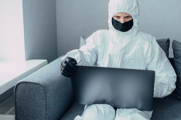 Medic in face mask and viral protective suit works online using a laptop and search news about coronavirus pandemic. Quarantined and self-isolated at home in curfews time and closed border.