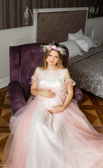 A pregnant woman in a pink dress sits on a soft chair in the bedroom. Waiting for a miracle.