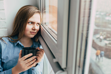 Caucasian teenage girl sits on windowsill, looks thoughtfully at the street outside the window and holds a black cup in her hands and is sad. In depression because of loneliness. self isolation time