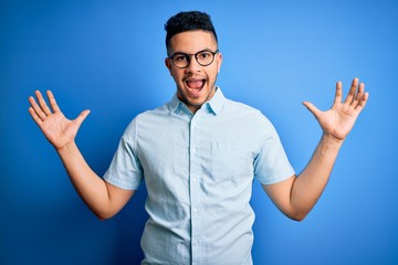Fototapeta premium Young handsome man wearing casual summer shirt and glasses over isolated blue background celebrating crazy and amazed for success with arms raised and open eyes screaming excited. Winner concept