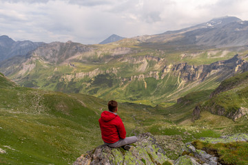 Fototapeta premium Young man in a red jacket on the stones looks at a magnificent view of the Alps mountains.
