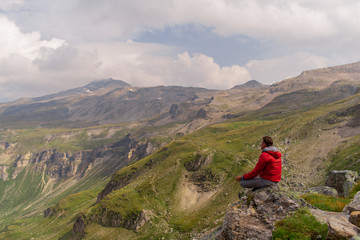 Young man in a red jacket on the stones looks at a magnificent view of the Alps mountains.