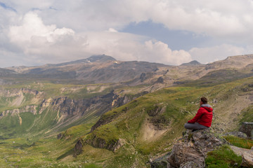 Young man in a red jacket on the stones looks at a magnificent view of the Alps mountains.