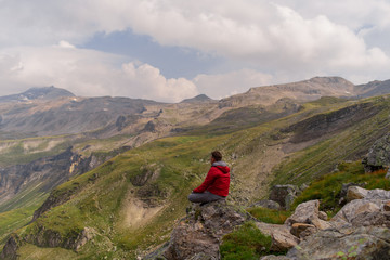 Young man in a red jacket on the stones looks at a magnificent view of the Alps mountains.