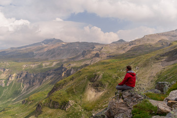 Naklejka premium Young man in a red jacket on the stones looks at a magnificent view of the Alps mountains.