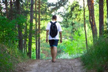 A guy in a white T-shirt, dark shorts and with a backpack walks a sand trail in the forest on a sunny summer day.
