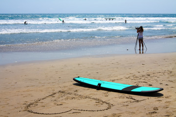 A surfboard is lying on the beach. Next to it, a large heart is drawn in the sand. Good weather, small waves. People learn to surf in the ocean.