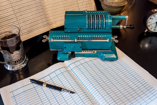 An old mechanical calculator, an arithmetic counter, accounting books, a watch, and a pen on a black table.