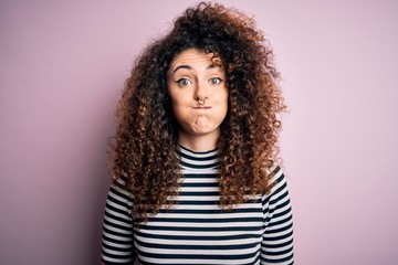 Young beautiful woman with curly hair and piercing wearing casual striped t-shirt puffing cheeks with funny face. Mouth inflated with air, crazy expression.