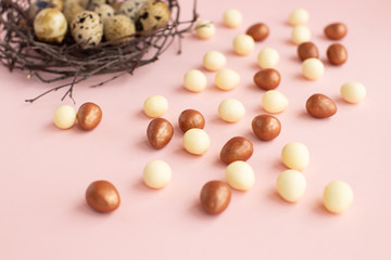 Nest of twigs with quail eggs on light pink background with small eggs made of white, dark chocolate. Easter greeting card, symbol of celebration Sunday of Christ. Holiday composition,congratulations