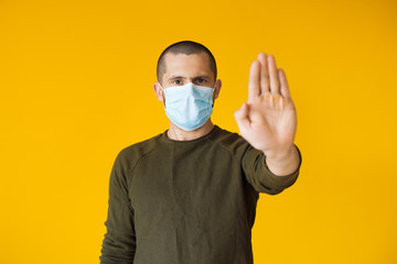 Caucasian man with short hair posing on a yellow wall is gesturing stop while wearing a special white mask