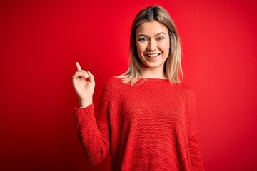 Young beautiful blonde woman wearing casual sweater over red isolated background with a big smile on face, pointing with hand finger to the side looking at the camera.