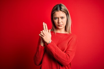 Young beautiful blonde woman wearing casual sweater over red isolated background Holding symbolic gun with hand gesture, playing killing shooting weapons, angry face