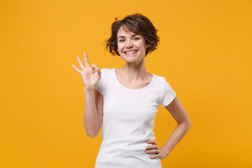 Smiling young brunette woman girl in white t-shirt posing isolated on yellow orange wall background studio portrait. People sincere emotions lifestyle concept. Mock up copy space. Showing OK gesture.